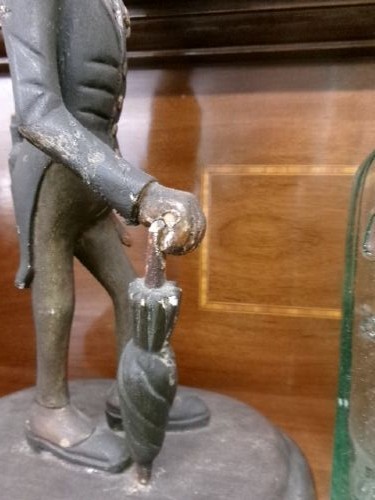 A carved and painted wooden figure of a caricatured man wearing a top hat and tailcoat, holding an umbrella while standing on a rounded grey base. View VII