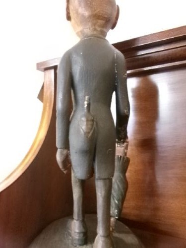 A carved and painted wooden figure of a caricatured man wearing a top hat and tailcoat, holding an umbrella while standing on a rounded grey base. View XII