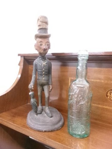 A carved and painted wooden figure of a caricatured man wearing a top hat and tailcoat, holding an umbrella while standing on a rounded grey base. View X