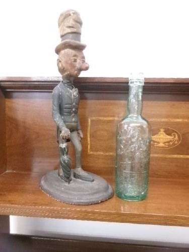 A carved and painted wooden figure of a caricatured man wearing a top hat and tailcoat, holding an umbrella while standing on a rounded grey base. View VIII
