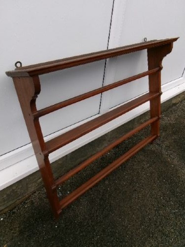 A mahogany wall-hanging rack featuring a top shelf, open tiers with horizontal retaining bars, shaped side brackets, and top brass mounting rings.