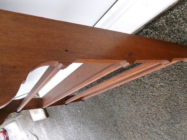 A mahogany wall-hanging rack featuring a top shelf, open tiers with horizontal retaining bars, shaped side brackets, and top brass mounting rings. View V
