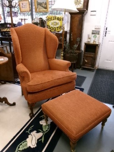 A high-backed Georgian style wing armchair with rolled arms, upholstered in textured orange fabric, accompanied by a matching square footstool.