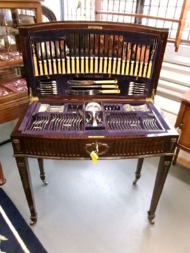 A dark mahogany freestanding cutlery cabinet with a open hinged lid displaying a fitted purple velvet interior with silver-plated flatware.