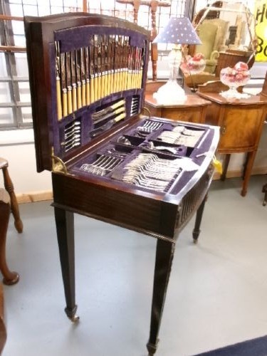 A dark mahogany freestanding cutlery cabinet with a open hinged lid displaying a fitted purple velvet interior with silver-plated flatware. View X
