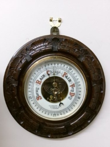 A circular Edwardian aneroid barometer set within a hand-carved mahogany frame with a brass bezel and visible mechanical movement.