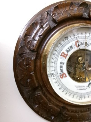 A circular Edwardian aneroid barometer set within a hand-carved mahogany frame with a brass bezel and visible mechanical movement. View II