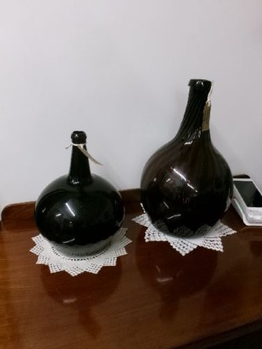 A dark green hand-blown glass onion bottle featuring a broad bulbous body, a long tapered neck, and an applied glass string rim. View VI