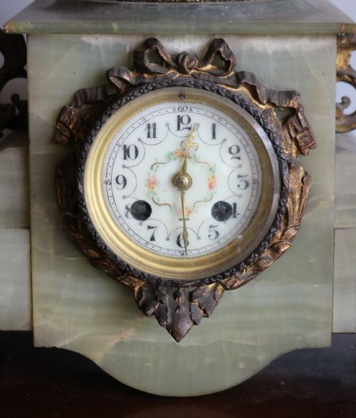 fig 1. An Art Nouveau French mantel clock featuring a bronzed female figure standing atop a light green onyx base with ornate brass feet and a decorated circular dial.