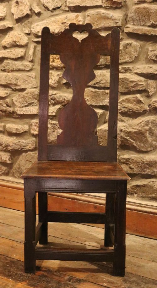 fig 1. An 18th-century dark oak Welsh backstool featuring a pierced heart cutout on the top rail, a vase-shaped splat back, and a solid panel seat supported by square stretchered legs.