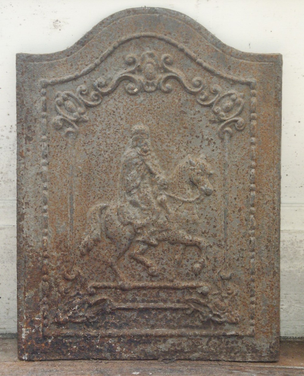 An arched cast iron fireback featuring an equestrian figure carved in raised relief surrounded by a scrolled decorative border with a rusted surface finish.