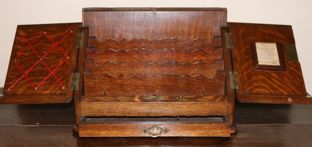 An open oak desktop stationery cabinet featuring tiered scalloped letter dividers, two hinged doors with ribbon memo webbing and frame fittings, and a single lower drawer.