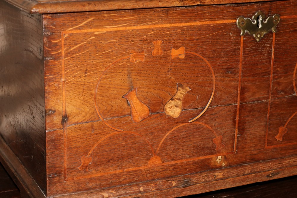 An 18th-century Welsh dark oak coffer bach featuring inlaid decorative scrolls on the front panel, bracket feet, and a central brass keyhole escutcheon. View V