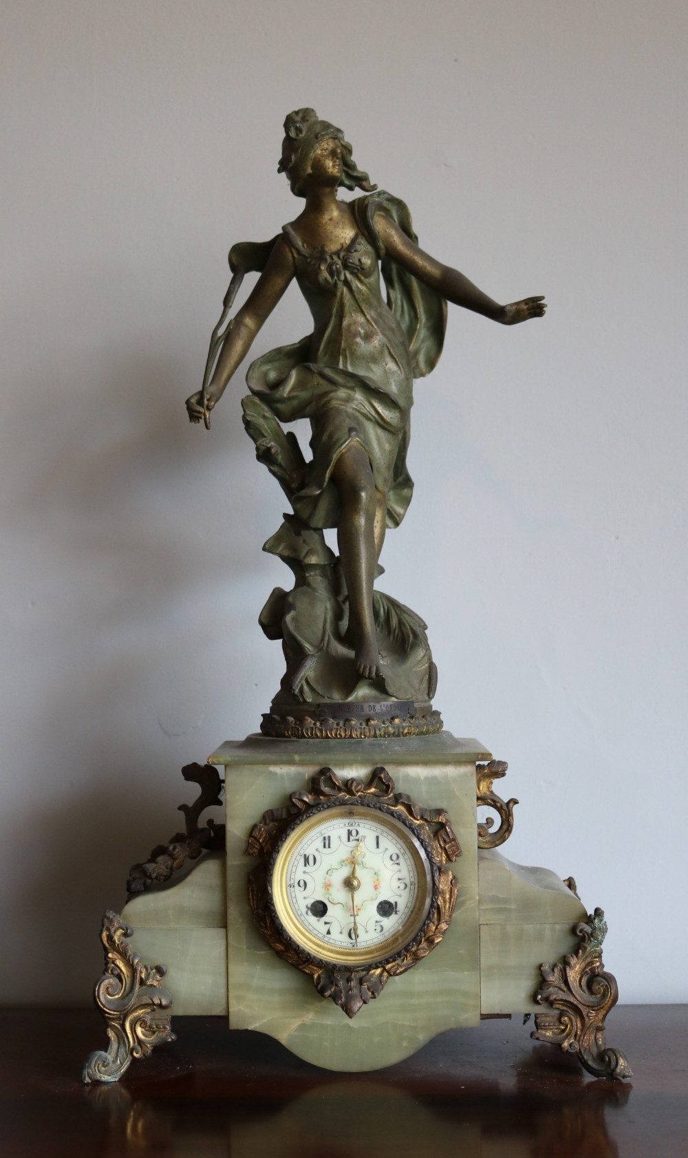 An Art Nouveau French mantel clock featuring a bronzed female figure standing atop a light green onyx base with ornate brass feet and a decorated circular dial.