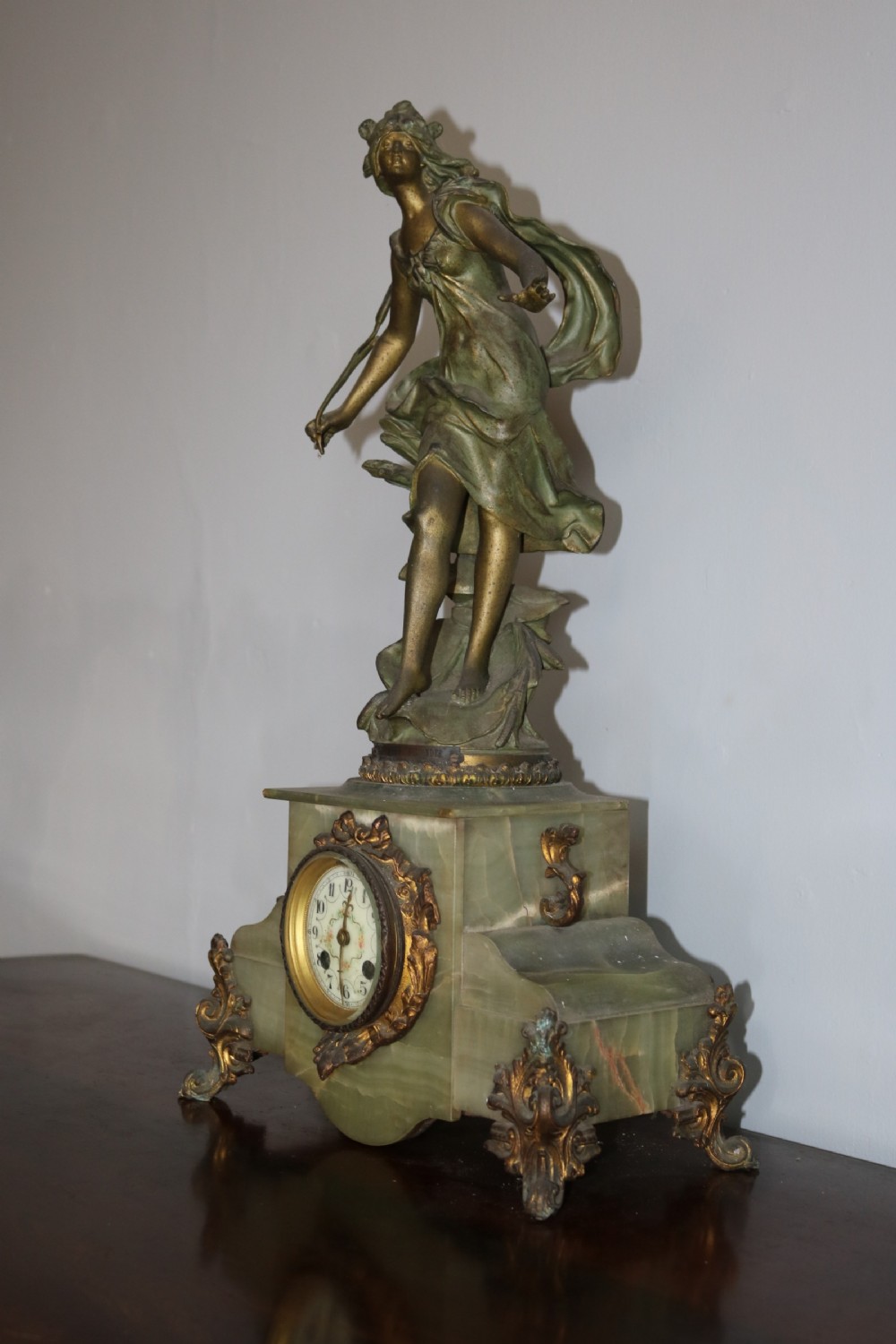 An Art Nouveau French mantel clock featuring a bronzed female figure standing atop a light green onyx base with ornate brass feet and a decorated circular dial. View V