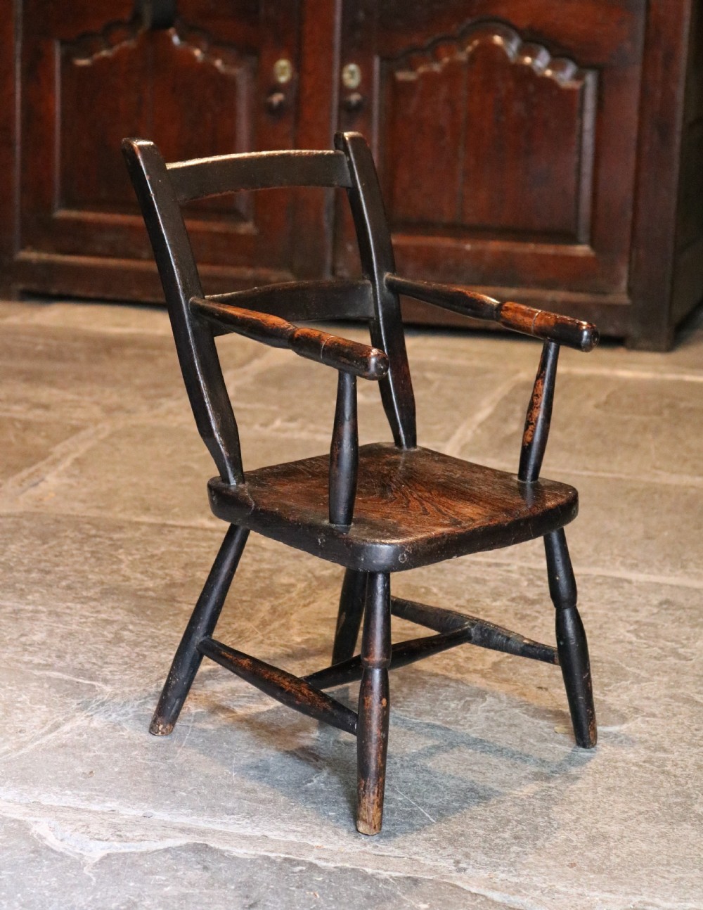 A nineteenth-century dark-stained Welsh childs primitive Windsor armchair featuring a thick slab seat, turned arms, and splayed legs.