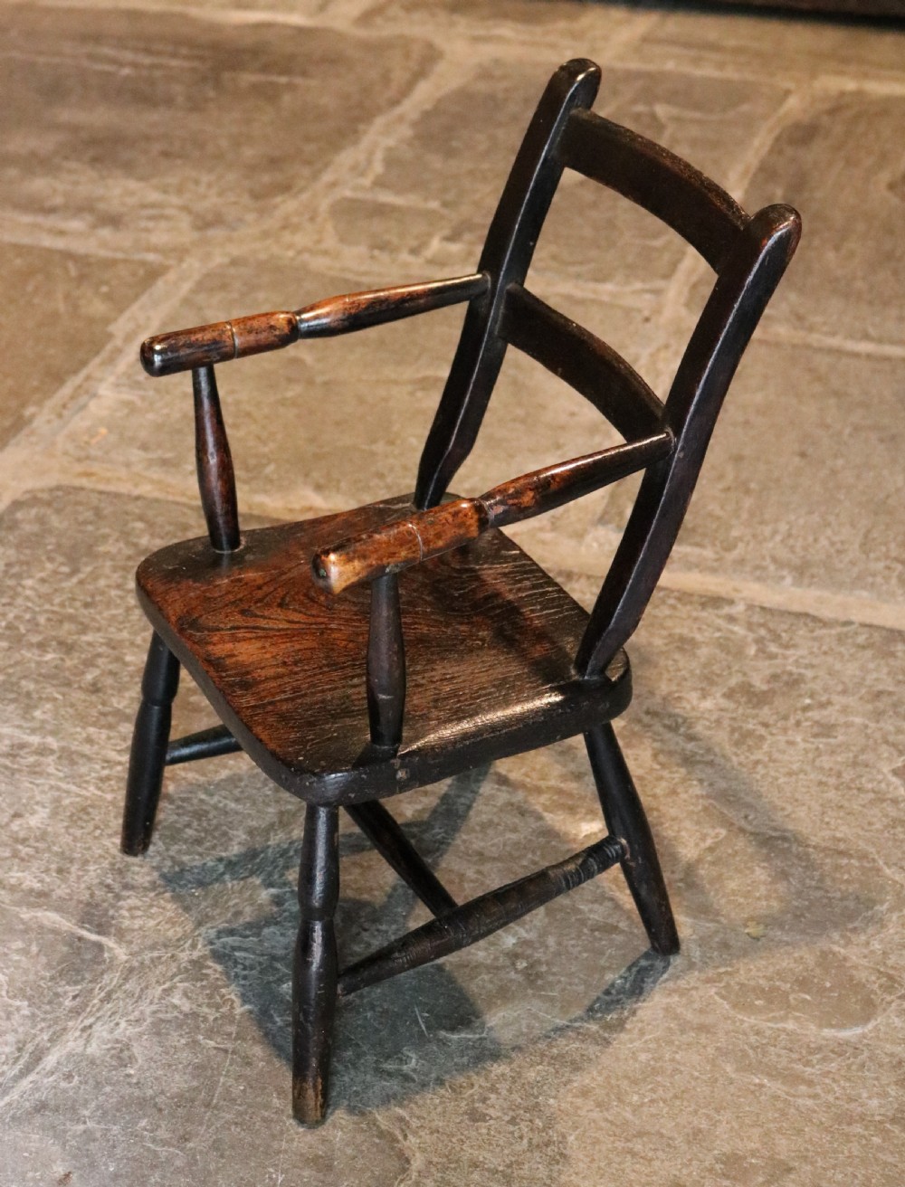 A nineteenth-century dark-stained Welsh childs primitive Windsor armchair featuring a thick slab seat, turned arms, and splayed legs. View V