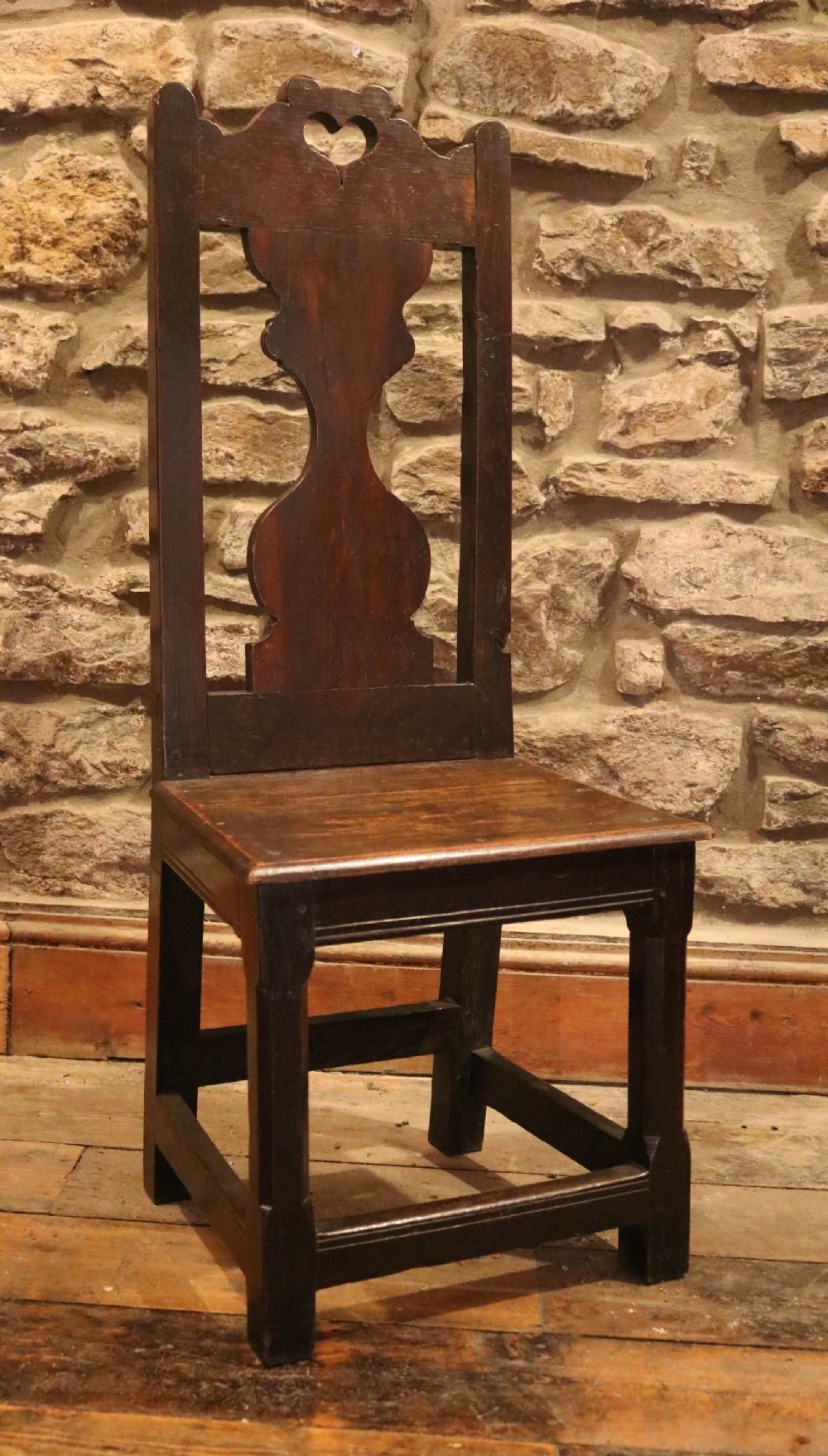 An 18th-century dark oak Welsh backstool featuring a pierced heart cutout on the top rail, a vase-shaped splat back, and a solid panel seat supported by square stretchered legs.