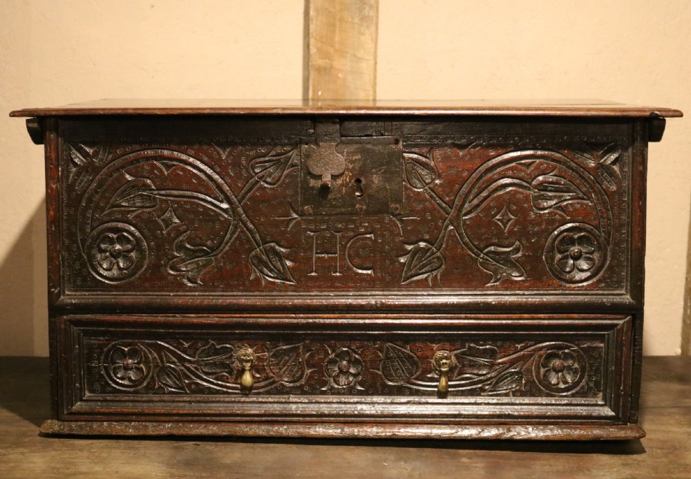 A 17th-century dark oak carved table box featuring scrollwork leaf motifs, carved initials HC, an iron lockplate, and a lower drawer.