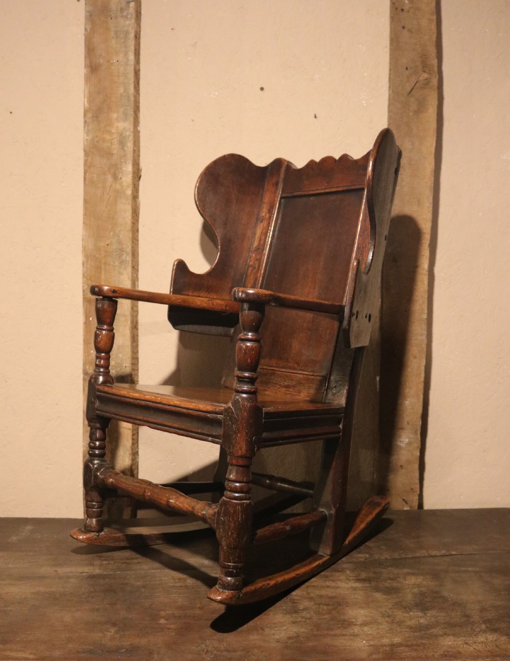 An antique dark oak childs lambing rocking chair featuring shaped winged sides, a panelled backrest, turned front supports, and curved wooden rockers. View II
