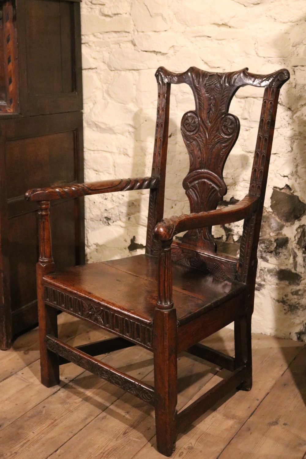 A dark carved oak Welsh armchair dating to the late 18th century with detailed foliate relief carving on the splat back and turned arm supports.
