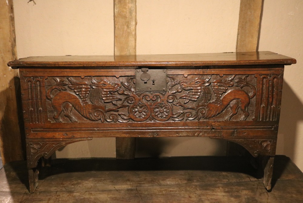 A dark brown carved oak chest featuring relief carvings of dragons, floral rosettes, and a central iron lock plate.