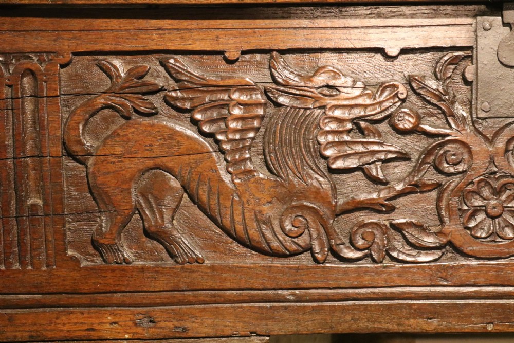 A dark brown carved oak chest featuring relief carvings of dragons, floral rosettes, and a central iron lock plate. View III