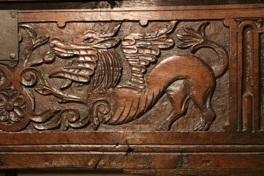 A dark brown carved oak chest featuring relief carvings of dragons, floral rosettes, and a central iron lock plate. View V