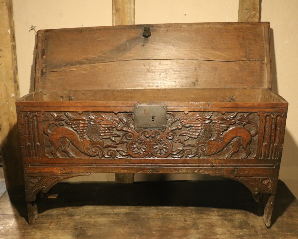 A dark brown carved oak chest featuring relief carvings of dragons, floral rosettes, and a central iron lock plate. View X
