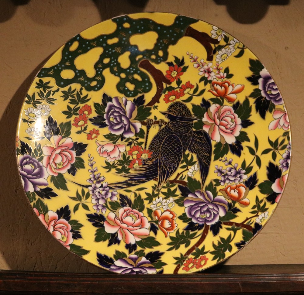 A circular antique ceramic charger with a bright yellow background decorated with colourful hand-painted peony flowers and a bird perched on a branch.