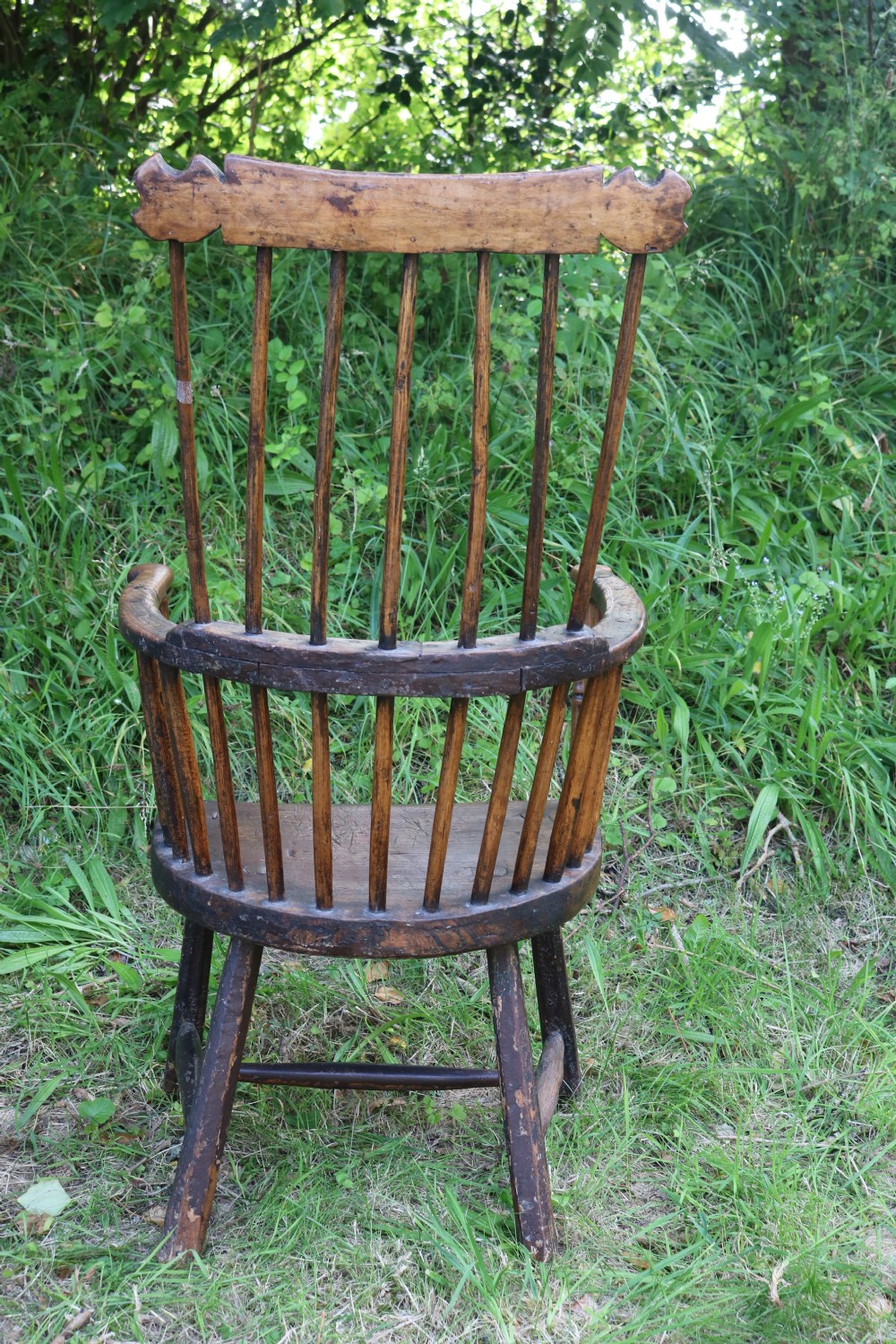 An 18th-century dark oak Welsh stick armchair featuring a shaped comb back, spindle uprights, a solid slab seat, and splayed legs. View V
