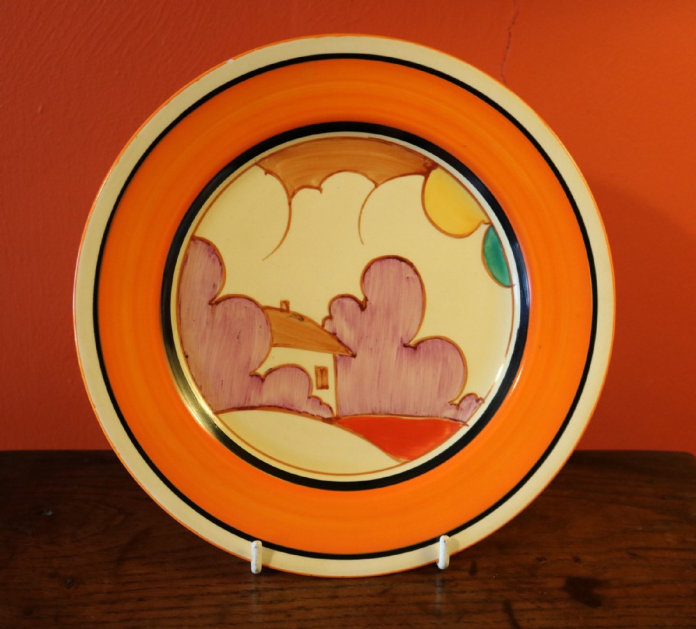 An Art Deco hand-painted ceramic plate featuring a stylised landscape scene with a cottage, purple trees, and a broad orange border.