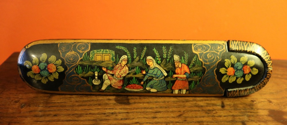 An elongated, rounded antique Indo-Persian lacquered papier-mache pen case featuring hand-painted figures in a landscape with floral end caps.