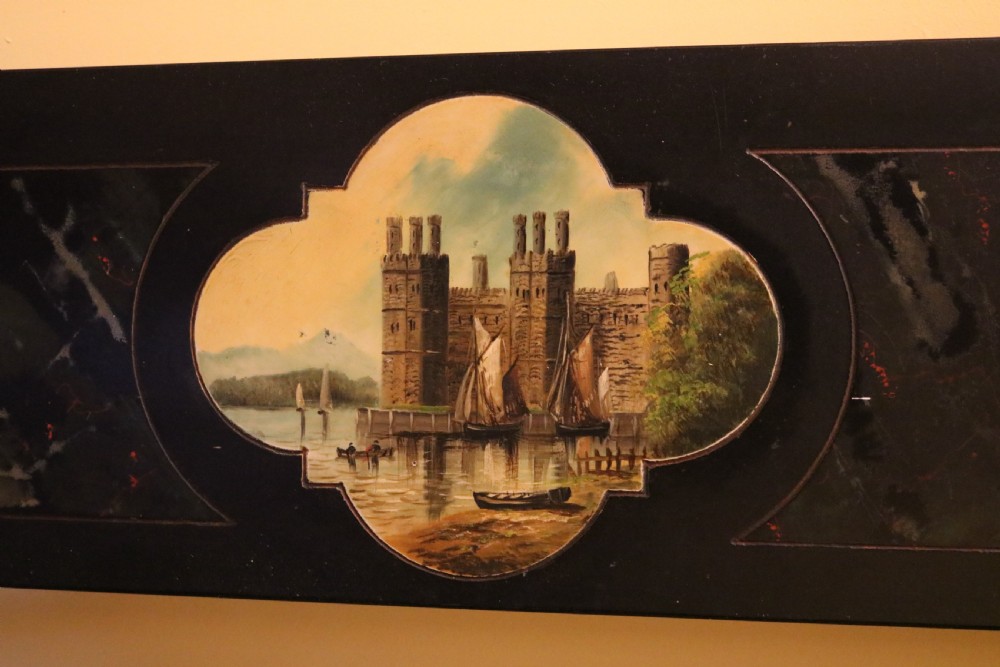 A long rectangular black slate panel featuring a central painted vignette of Caernarfon Castle within a quatrefoil border and faux-marbled side sections. View IV