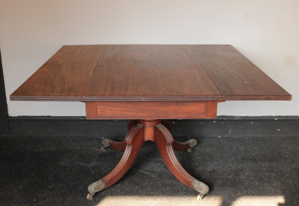 A Regency mahogany dining table featuring a rectangular drop-leaf top supported by a central reeded pedestal and four splayed legs with brass toe castors.