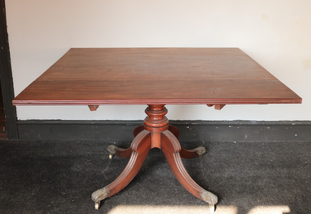 A Regency mahogany dining table featuring a rectangular drop-leaf top supported by a central reeded pedestal and four splayed legs with brass toe castors. View II