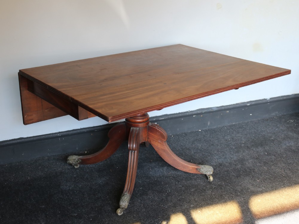 A Regency mahogany dining table featuring a rectangular drop-leaf top supported by a central reeded pedestal and four splayed legs with brass toe castors. View IV