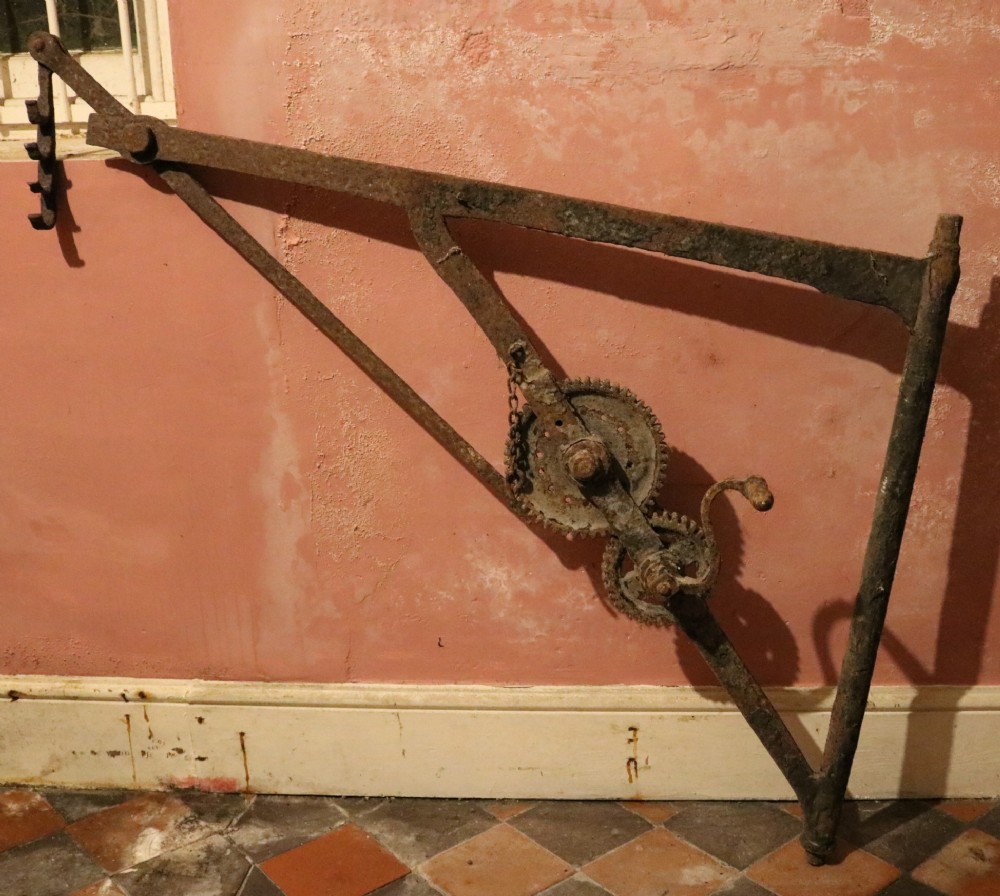 An eighteenth-century wrought iron hearth crane featuring a gear mechanism, hand crank, triangular bracing, and notched pot hook.