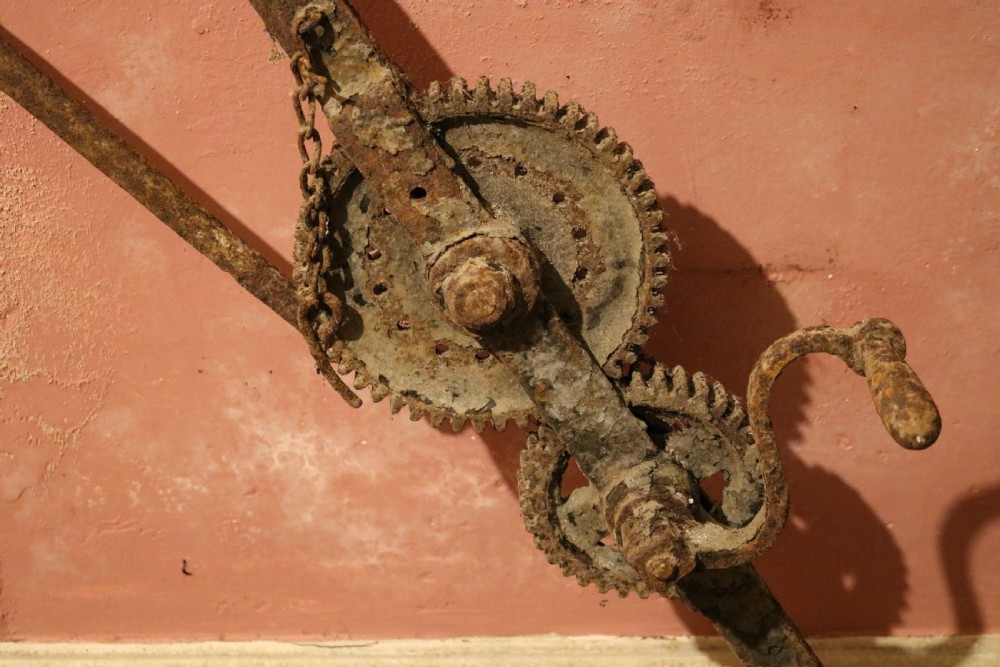 An eighteenth-century wrought iron hearth crane featuring a gear mechanism, hand crank, triangular bracing, and notched pot hook. View II