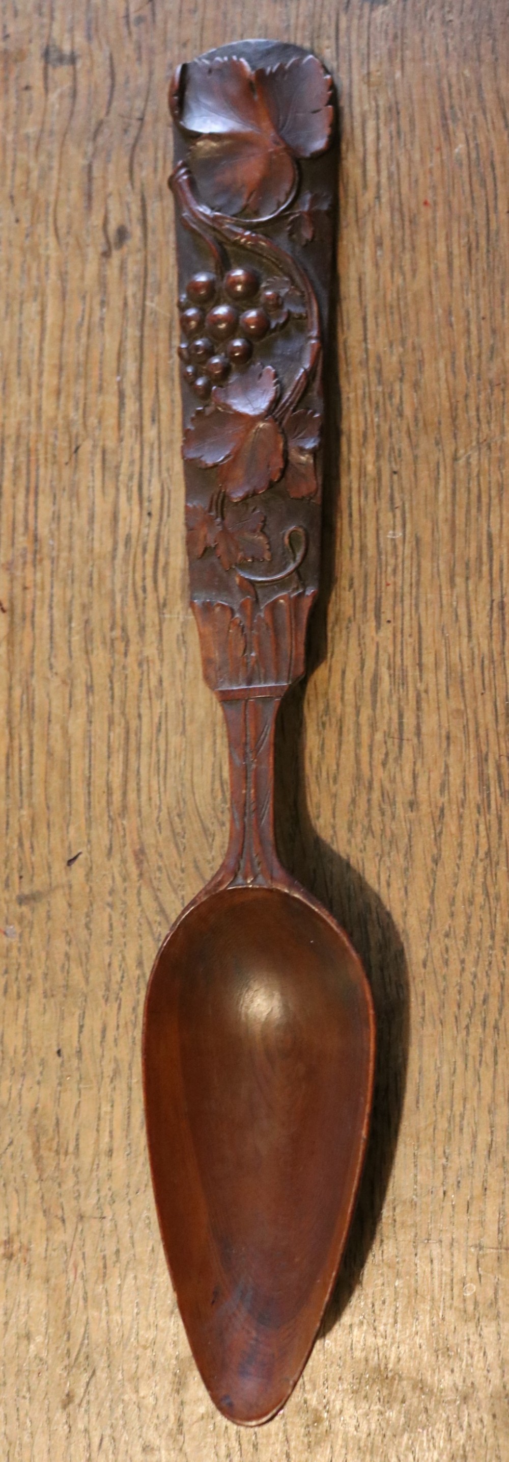 A hand-carved dark yew-wood spoon featuring a detailed relief design of grapevines and fruit along its flat handle.