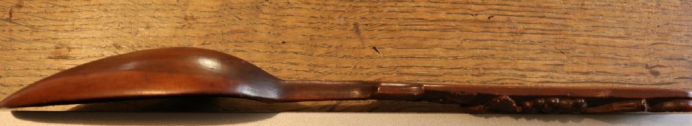 A hand-carved dark yew-wood spoon featuring a detailed relief design of grapevines and fruit along its flat handle. View V