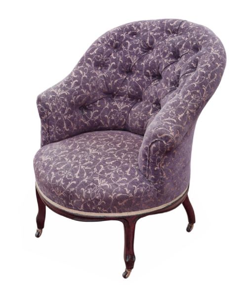 fig 1. An antique Victorian tub armchair upholstered in patterned dusty purple fabric with a button-tufted backrest and mahogany legs on brass castors.