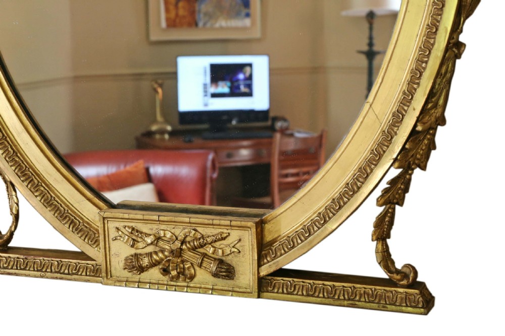 An antique gold gilt oval wall mirror featuring a Neoclassical ribbon motif at the base and foliage side supports. View VI