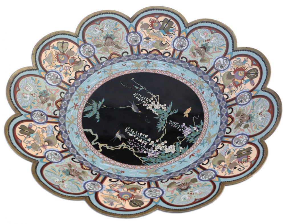 A large Japanese Meiji period scalloped cloisonne charger plate featuring a black central panel decorated with wisteria and birds, framed by intricate polychrome enamel outer borders.