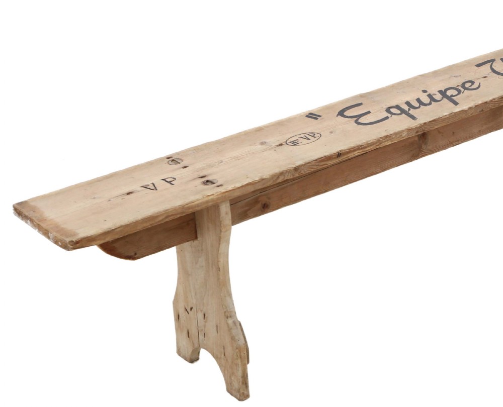 A long, pale timber garden bench featuring stencilled black text along the top plank and shaped trestle legs. View II