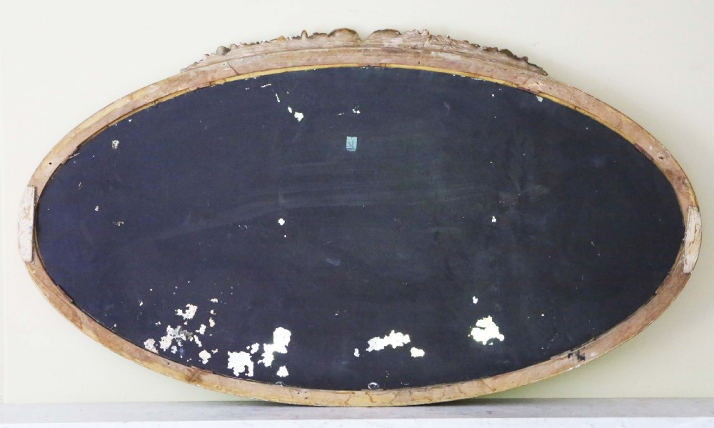 A large horizontal oval wall mirror with a gilded frame topped by a carved floral cresting and featuring foxed glass. View VI