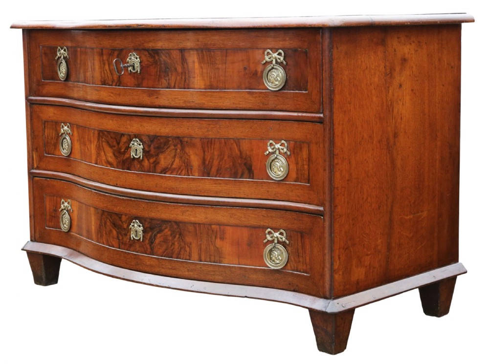 An 18th-century Continental oak and walnut chest of drawers featuring a serpentine front, crossbanded top, and three drawers with ornamental brass handles. View IV