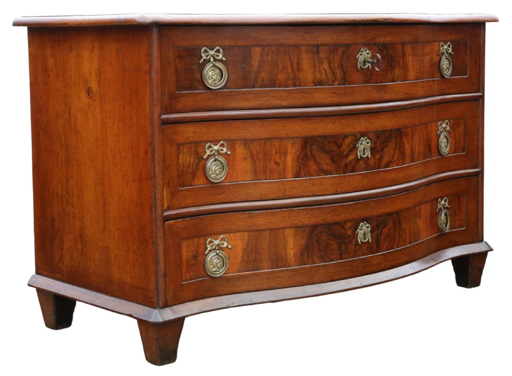 An 18th-century Continental oak and walnut chest of drawers featuring a serpentine front, crossbanded top, and three drawers with ornamental brass handles. View V