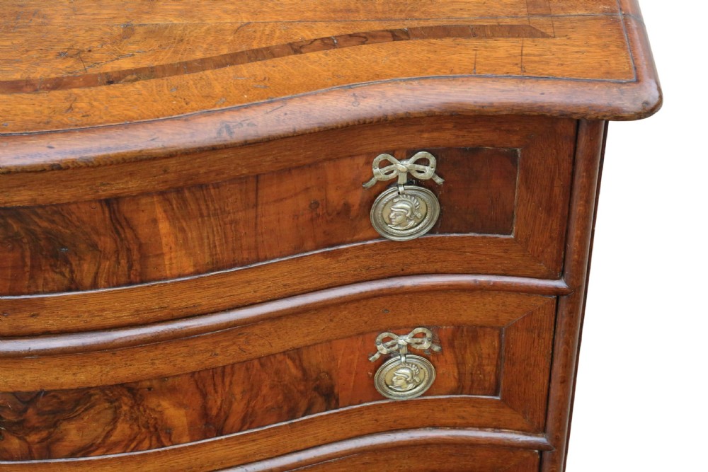 An 18th-century Continental oak and walnut chest of drawers featuring a serpentine front, crossbanded top, and three drawers with ornamental brass handles. View VII