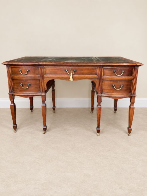 fig 1. Late nineteenth-century mahogany writing desk featuring an inset dark leather top, five drawers with brass swan-neck handles, fluted pilasters, and tapered legs with brass castors.